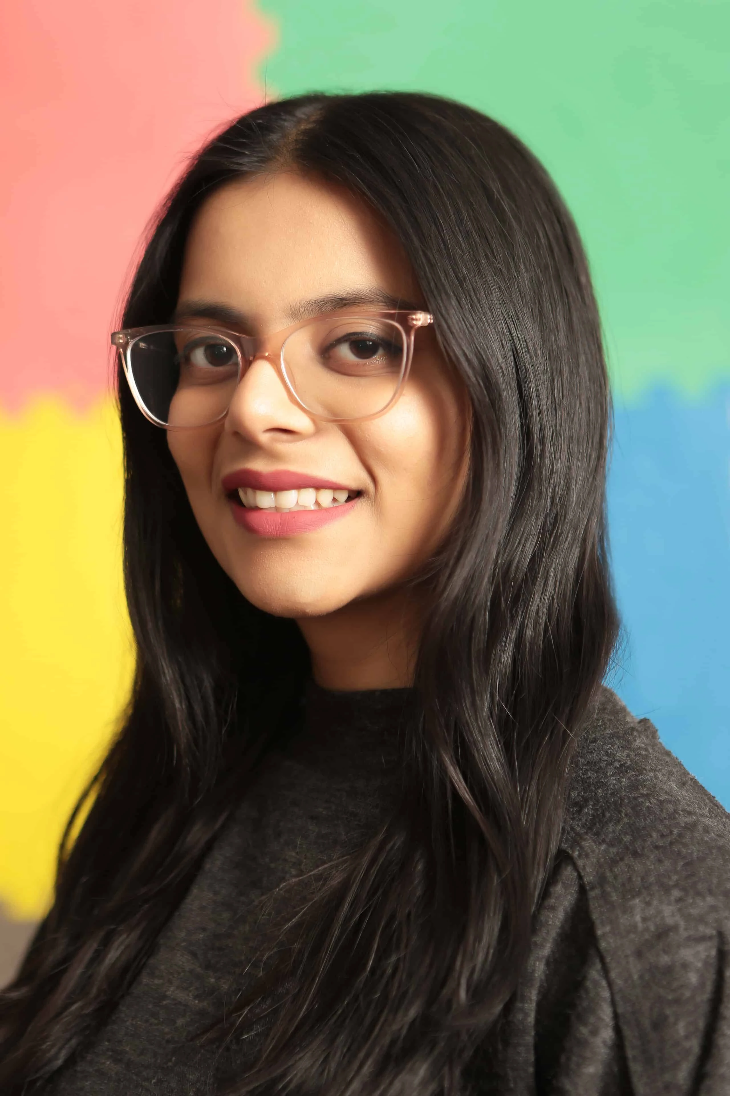 Neha wearing glasses and smiling in front of a bright, playful backdrop